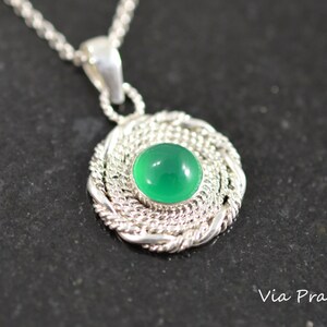May include: A silver pendant necklace featuring a round, bright green gemstone set in a woven silver frame. The pendant hangs from a delicate silver chain. The necklace is displayed on a dark surface.