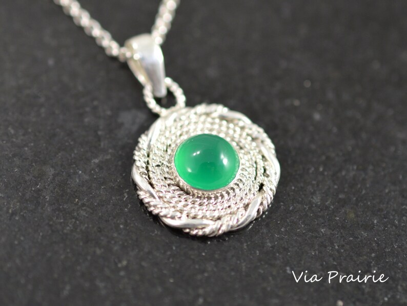 May include: A silver pendant necklace featuring a round, bright green gemstone set in a woven silver frame. The pendant hangs from a delicate silver chain. The necklace is displayed on a dark surface.