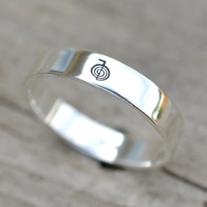 Puede incluir: Un anillo de banda de plata con un s&iacute;mbolo grabado simple en la parte superior.