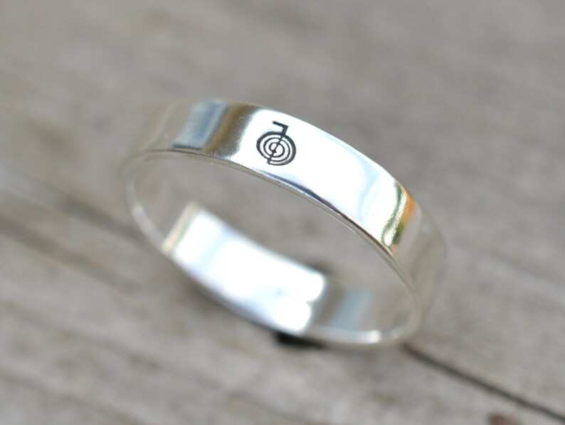 Puede incluir: Un anillo de banda de plata con un s&iacute;mbolo grabado simple en la parte superior.