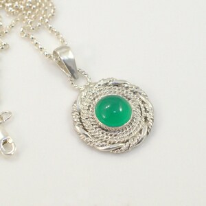 May include: A silver necklace with a round pendant featuring a vibrant green gemstone. The pendant is encircled by a twisted rope design. The necklace chain is a delicate silver ball chain.
