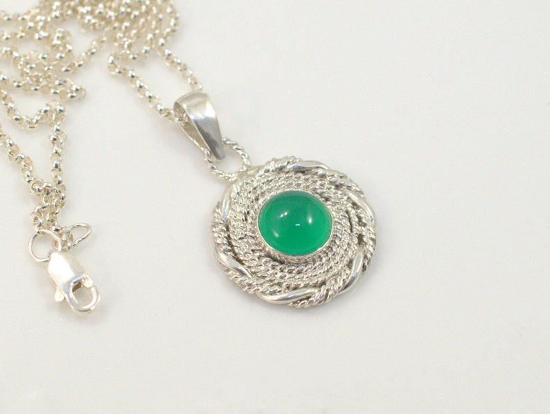 May include: A silver necklace with a round pendant featuring a vibrant green gemstone. The pendant is encircled by a twisted rope design. The necklace chain is a delicate silver ball chain.