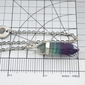 Puede incluir: Una cadena de plata con un colgante de plata que presenta una punta de cristal de fluorita verde, azul y p&uacute;rpura. El colgante tambi&eacute;n tiene un dije de plata con un s&iacute;mbolo en espiral.