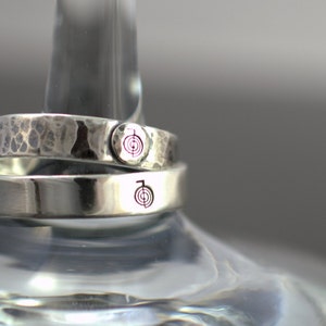 Puede incluir: Dos anillos de plata con textura martillada. El anillo superior tiene un peque&ntilde;o s&iacute;mbolo grabado en el lateral. El anillo inferior tiene un s&iacute;mbolo grabado m&aacute;s grande en el lateral.