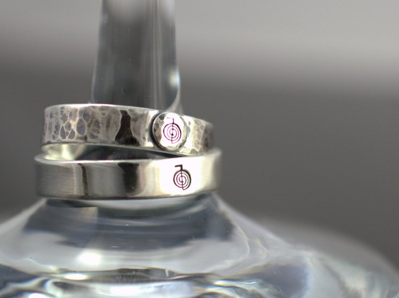 Puede incluir: Dos anillos de plata con textura martillada. El anillo superior tiene un peque&ntilde;o s&iacute;mbolo grabado en el lateral. El anillo inferior tiene un s&iacute;mbolo grabado m&aacute;s grande en el lateral.