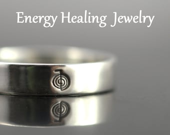 May include: Silver ring with a symbol engraved on the band. The symbol is a circle with a line through it, and a smaller circle inside the larger circle. The ring is on a gray surface.