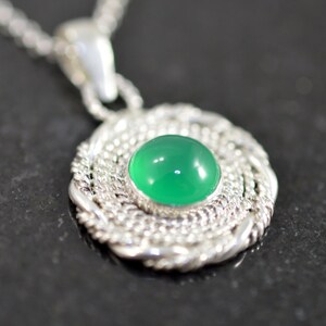 May include: A silver pendant necklace featuring a round, bright green gemstone set in a detailed, woven silver frame. The pendant hangs from a delicate silver chain, creating a classic and elegant jewelry piece.