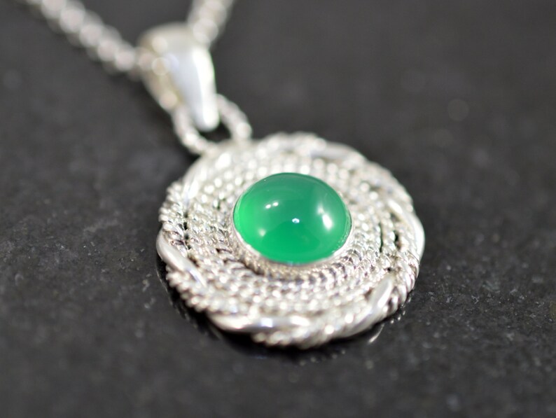May include: A silver pendant necklace featuring a round, bright green gemstone set in a detailed, woven silver frame. The pendant hangs from a delicate silver chain, creating a classic and elegant jewelry piece.