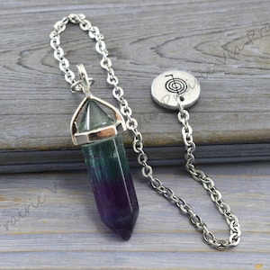 Puede incluir: Un p&eacute;ndulo de cristal de fluorita verde, morado y azul con una cadena de plata y un dije de plata con un s&iacute;mbolo.