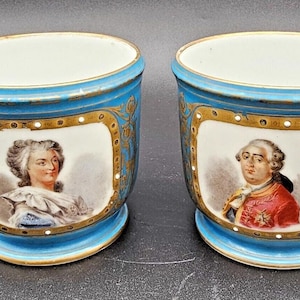 May include: Pair of blue and white porcelain cups with gold trim. Each cup features a portrait of a person in a gold frame.
