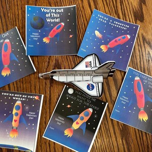 Puede incluir: Un conjunto de diez tarjetas de San Valent&iacute;n con tem&aacute;tica espacial que presentan cohetes, planetas y estrellas. Las tarjetas tienen un fondo azul con texto blanco y est&aacute;n impresas con la frase "You're out of this world!" y otros juegos de palabras con tem&aacute;tica espacial.