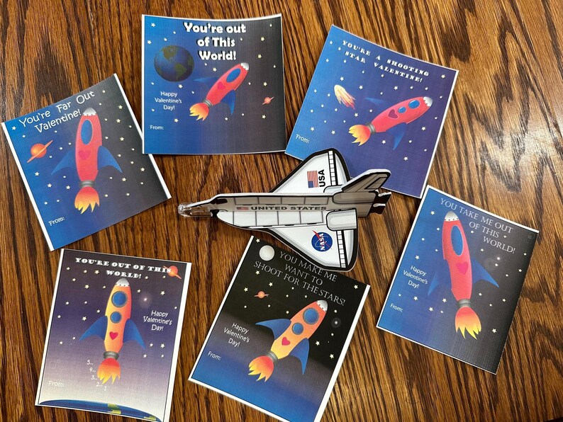 Puede incluir: Un conjunto de diez tarjetas de San Valent&iacute;n con tem&aacute;tica espacial que presentan cohetes, planetas y estrellas. Las tarjetas tienen un fondo azul con texto blanco y est&aacute;n impresas con la frase "You're out of this world!" y otros juegos de palabras con tem&aacute;tica espacial.