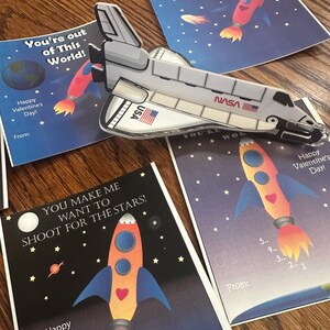 Puede incluir: Cuatro tarjetas coloridas del D&iacute;a de San Valent&iacute;n con ilustraciones de cohetes y con tem&aacute;tica espacial. Las tarjetas tienen el texto "You're out of this world!" y "You make me want to shoot for the stars!"