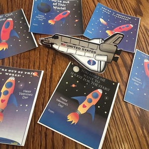Puede incluir: Un conjunto de diez tarjetas de San Valent&iacute;n con tem&aacute;tica espacial. Las tarjetas tienen un fondo azul con estrellas y planetas. Cada tarjeta presenta una nave espacial y un mensaje diferente para el D&iacute;a de San Valent&iacute;n. Los mensajes incluyen "You're Far Out Valentine!" y "You Make Me Want To Shoot For The Stars!"