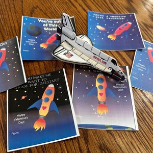 Puede incluir: Cinco tarjetas del D&iacute;a de San Valent&iacute;n con tem&aacute;tica espacial. Las tarjetas muestran cohetes, estrellas y el texto "You're Far Out Valentine!"  "You're out of This World"  "You Make Me Want To Shoot For The Stars!"  "You'll Wonder"  "You Take Me Out Of This World!"