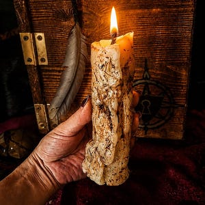May include: A tall, cylindrical pillar candle with a textured, melted wax surface embedded with dried herbs and botanical elements. The candle features a single lit wick with a bright orange flame, casting a warm glow over the uneven, cream-colored wax.