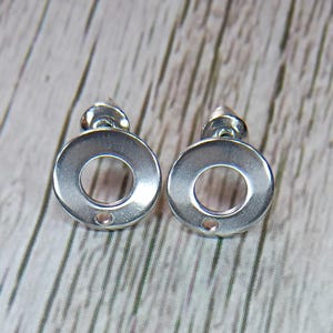May include: A pair of silver-toned metal stud earrings featuring a circular design with a central cutout and a small hole at the bottom edge for attaching additional components. The earrings have a smooth, reflective finish and include post backs.