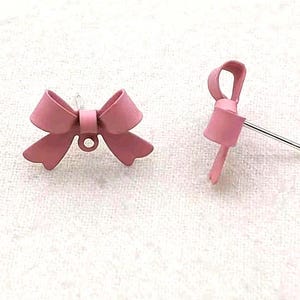 May include: A pair of pink bow-shaped stud earrings. One earring is a complete bow with a post and a small loop. The other earring is a bow with a post. The earrings are a solid, matte pink color.