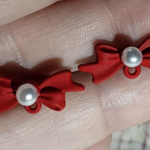 May include: Two red bow-shaped ornaments, each with a white pearl center and a small loop. The bows are a vibrant red color, and the pearls are a bright white. These decorative elements could be used for jewelry or craft projects.