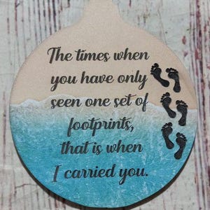 May include: A round wooden ornament with a blue and white beach scene and the text "The times when you have only seen one set of footprints, that is when I carried you."