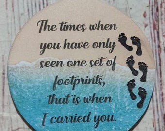 Peut inclure: Une décoration ronde en bois avec une scène de plage bleue et blanche et le texte "The times when you have only seen one set of footprints, that is when I carried you."