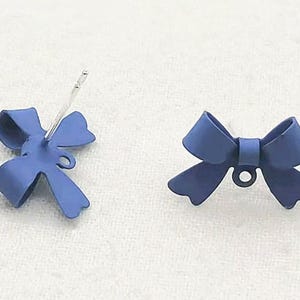 May include: Two blue bow-shaped earrings. One earring has a silver post, and the other is a bow with a small loop. The earrings are a solid, matte blue color. The background is a white, textured surface.