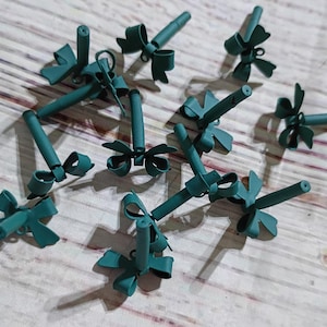 May include: A collection of teal-colored floral stem holders with bow accents. The holders are cylindrical with a bow at the base and a small opening at the top. They are arranged on a light wood-grain surface.