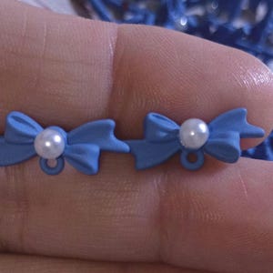 May include: Two light blue bow-shaped embellishments with a white pearl in the center. Each bow has a small loop at the bottom. The bows are held in a hand, with blue decorative elements in the background.