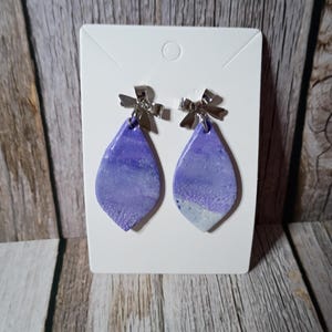 May include: A pair of teardrop-shaped earrings featuring a marbled purple and white textured finish. Each earring is attached to a silver-toned metal bow accent at the top, mounted on a white rectangular display card with a circular hole at the top.