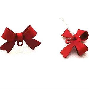 May include: Two red bow-shaped ornaments. One bow is shown from the front, and the other is shown from the side with a silver wire attached. The bows have a loop for hanging. The background is white.