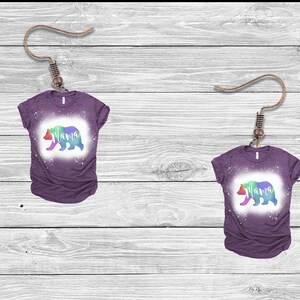 May include: A pair of purple t-shirts with a rainbow bear silhouette and the word "mama" printed on them. The shirts are hanging from earring hooks.