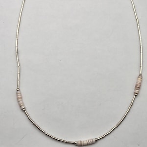 以下が含まれることがあります： 小さな円筒形のビーズと、より大きな薄いピンクと白の貝殻ビーズが交互に配置された、繊細なシルバーのネックレス。 ネックレスはシンプルでミニマリストなデザインです。 ネックレスの長さは約45cmです。