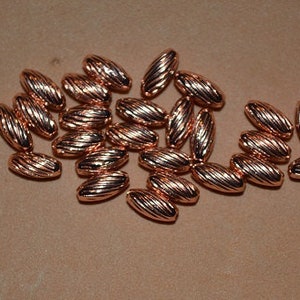 Puede incluir: Un grupo de aproximadamente 30 cuentas ovaladas de color cobre con una textura acanalada. Las cuentas están dispersas sobre una superficie marrón.