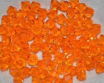 Puede incluir: Un gran grupo de pequeñas cuentas de vidrio facetadas de color naranja. Las cuentas están dispuestas en un montón suelto sobre una superficie blanca.