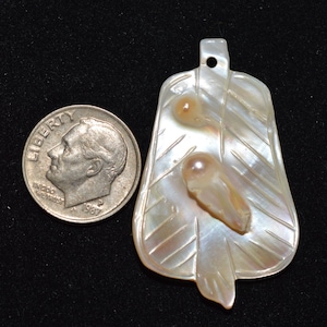 May include: A mother-of-pearl leaf pendant with two embedded, light-coloured, irregular-shaped stones. The pendant has a small hole at the top. A U.S. dime is next to the pendant for size comparison.
