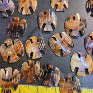 May include: A collection of 15 brown and white carved eagle-shaped pendants made from shell. The pendants are approximately 2 inches in length and feature intricate details.