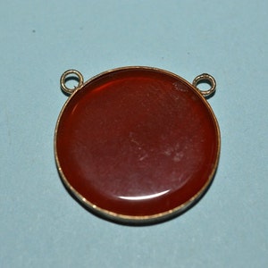 Pode incluir: Um pingente redondo de pedra vermelha com uma configuração de metal dourado e dois laços para prender uma corrente.