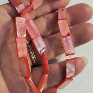 May include: A necklace composed of rectangular, coral-colored beads with a pearlescent sheen. The beads are strung together, creating a flexible strand. The necklace is held in a hand, showcasing its color and design.