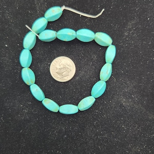 Puede incluir: Una hebra de cuentas ovaladas de color turquesa, aproximadamente del tamaño de una moneda de diez céntimos de euro, enhebradas en un cordón blanco.