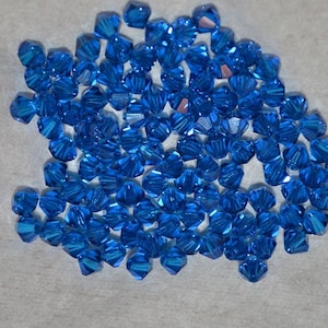 May include: A large group of small, faceted, blue glass beads. The beads are scattered on a white surface.