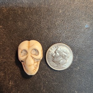 Puede incluir: Una cuenta de calavera blanca con detalles intrincados, junto a una moneda de diez centavos de dólar estadounidense para comparar el tamaño.