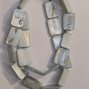 May include: A necklace composed of rectangular, iridescent, pale gray beads. The beads are strung together on a black cord, creating a layered effect. The necklace has a subtle sheen and a natural, organic appearance.
