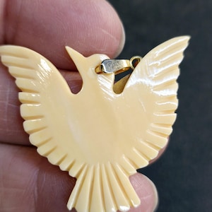 Puede incluir: Un colgante de pájaro tallado de color crema con una anilla de oro.