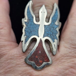 Puede incluir: Anillo plateado con diseño de ángel. Las alas del ángel están incrustadas con pequeños trozos de turquesa, y la parte inferior está incrustada con piedra roja. El anillo se lleva en un dedo.