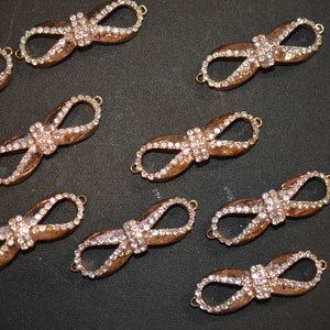 Puede incluir: Primer plano de varios hallazgos de joyería en forma de lazo de color bronce. Cada lazo está adornado con numerosas piedras de estrás pequeñas y transparentes. Los hallazgos tienen pequeños bucles en cada extremo para la fijación. El fondo es de color oscuro y liso.