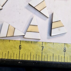 May include: A set of six white triangular tiles with gold and cream stripes. The tiles are arranged on a white surface with a yellow ruler in the foreground.