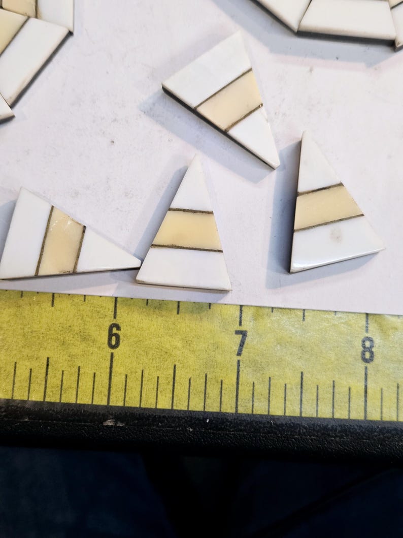May include: A set of six white triangular tiles with gold and cream stripes. The tiles are arranged on a white surface with a yellow ruler in the foreground.