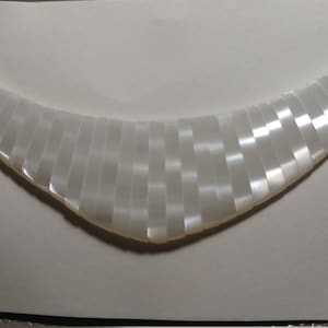 May include: A white iridescent necklace made of small, rectangular pieces of shell. The necklace is curved and has a slightly pointed shape.