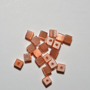 May include: A group of small, square, peach-colored beads with a shimmery finish. The beads have a hole in the center and are scattered on a white surface.