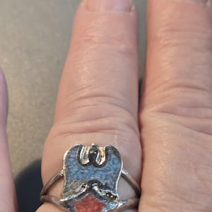 Puede incluir: Anillo plateado con un diseño en forma de escudo con incrustaciones en azul claro y rojo. El diseño del anillo incluye un emblema de águila o pájaro en la parte superior y una forma de diamante en la parte inferior. El anillo se lleva en un dedo.